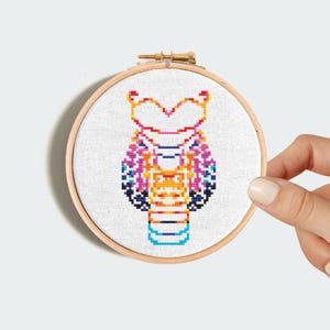 Puede incluir: Un bordado de punto de cruz colorido de un búho estilizado con una cabeza en forma de corazón, con una paleta de colores arcoíris sobre un fondo blanco.