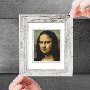 Puede incluir: Una impresión enmarcada de una pintura de la Mona Lisa. La pintura está en un marco blanco con un acabado de madera desgastada. La Mona Lisa está representada con una sonrisa suave y su cabello está recogido hacia atrás.