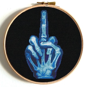 Puede incluir: Un bordado de punto de cruz en un aro de madera. El diseño presenta una mano esquelética azul y blanca haciendo la señal del dedo corazón sobre un fondo negro. El aro es redondo y tiene un cierre de latón.