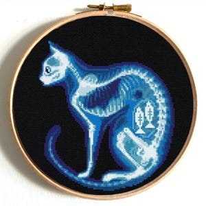 Puede incluir: Una bordado de punto de cruz azul y blanco de un esqueleto de gato sobre un fondo negro. El gato está sentado con la cola enroscada alrededor de su cuerpo.
