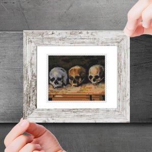 May include: A framed print of a still life painting depicting three human skulls on a wooden table. The skulls are painted in shades of grey, brown, and white.