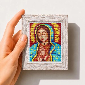 May include: A small cross stitch pattern featuring a depiction of the Virgin of Guadalupe with a red background, blue mantle, and yellow halo, framed in a white textured wooden frame, with the text Mini Art Cross Stitch Pattern PDF below.