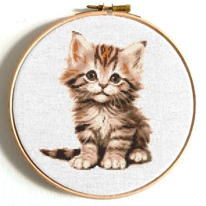 Könnte beinhalten: Eine Kreuzstich-Stickerei eines süßen, braun-weißen Tabby-Kätzchens, das mit gekreuzten Pfoten sitzt. Das Kätzchen hat große, grüne Augen und einen süßen Ausdruck.