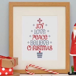 May include: Framed cross-stitch art with a Christmas tree design. The tree is composed of the words "JOY, LOVE, PEACE, BELIEVE, CHRISTMAS" in red and gray. A red Santa Claus figurine and wrapped gifts are also in the image.