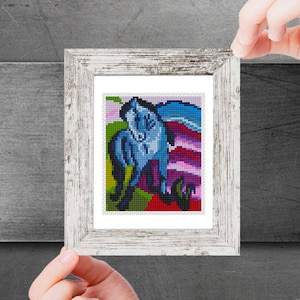 May include: A framed cross-stitch artwork depicting a blue horse with a pink and purple background. The horse is standing on a green field with flowers.