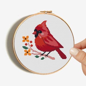 May include: Cross stitch pattern of a red cardinal bird perched on a branch with yellow flowers. The pattern is on white Aida cloth in a wooden embroidery hoop.