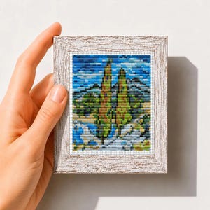 May include: A cross stitch pattern featuring a landscape with two tall cypress trees, rolling hills, and a blue sky, framed in a textured white wooden frame. The text "Mini Art Cross Stitch Pattern PDF" is printed at the bottom of the image.