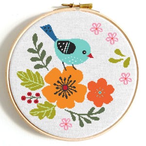 May include: A cross-stitch embroidery hoop with a colorful design of a blue bird perched on a flower. The flower is orange with a black center and there are green leaves and pink flowers surrounding the bird.