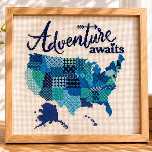 May include: Cross-stitch artwork in a wooden frame. The design features a map of the United States in shades of blue, with the words "Adventure awaits" stitched above. The artwork is displayed on a wooden surface.