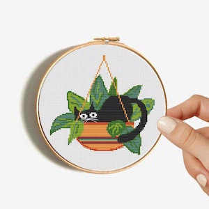May include: A cross-stitch embroidery hoop showing a black cat in a hanging plant pot. The pot is orange with green stripes, and the leaves are green. The cat has white eyes and a white mustache. The hoop is held by a hand.