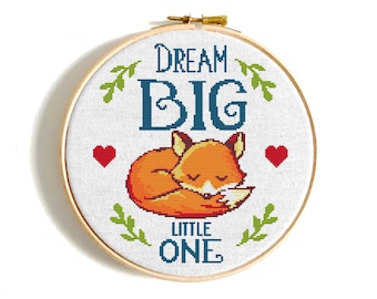 Op de afbeelding: Een kruissteek borduurring met een slapende oranje vos en de woorden "DREAM BIG LITTLE ONE" in blauw en wit. Twee rode harten en groene bladaccenten omringen de tekst en de vos.