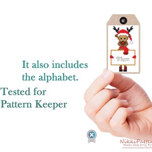 Puede incluir: Una etiqueta de regalo de punto de cruz con un reno con gorro y bufanda de Pap&aacute; Noel, sosteniendo un letrero que dice "Mum". La etiqueta es marr&oacute;n con un ojal negro. Tambi&eacute;n se ve el texto "It also includes the alphabet. Tested for Pattern Keeper".