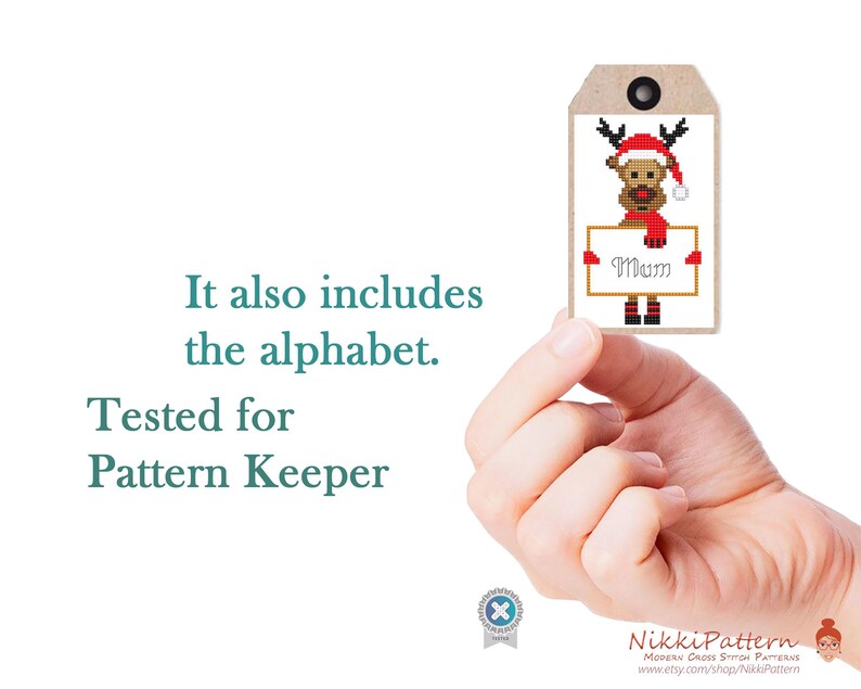 Puede incluir: Una etiqueta de regalo de punto de cruz con un reno con gorro y bufanda de Pap&aacute; Noel, sosteniendo un letrero que dice "Mum". La etiqueta es marr&oacute;n con un ojal negro. Tambi&eacute;n se ve el texto "It also includes the alphabet. Tested for Pattern Keeper".