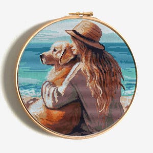 May include: Cross-stitch art in a wooden hoop depicts a person hugging a golden retriever on a beach. The person wears a hat and has long brown hair. The background shows the ocean and sky.