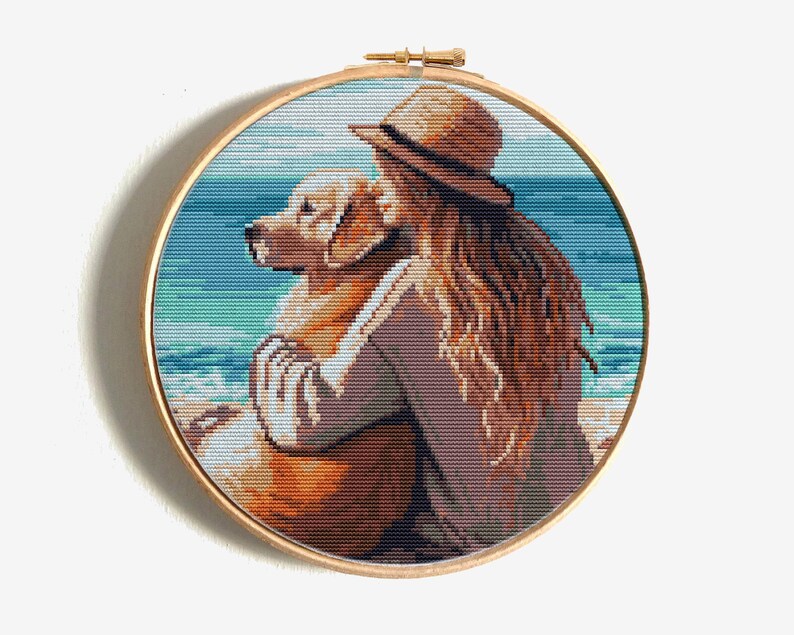 May include: Cross-stitch art in a wooden hoop depicts a person hugging a golden retriever on a beach. The person wears a hat and has long brown hair. The background shows the ocean and sky.