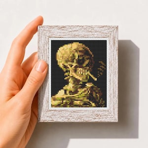 May include: A framed cross stitch pattern featuring a pixelated skull smoking a cigarette against a black background. The artwork is displayed in a textured white wooden frame. The text "Mini Art Cross Stitch Pattern PDF" is printed below.