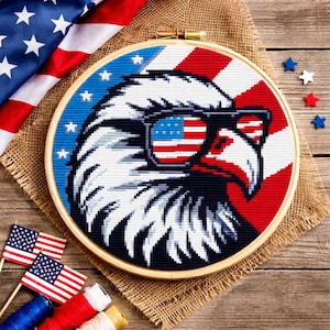 May include: A cross-stitch hoop art piece featuring an American bald eagle wearing sunglasses with the American flag design. The eagle is set against a blue and red background, with a wooden hoop. Patriotic theme.