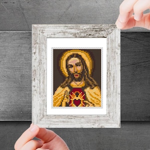 Puede incluir: Una imagen enmarcada de Jesucristo con un halo dorado y un corazón rojo en su pecho. La imagen está sobre un fondo oscuro.