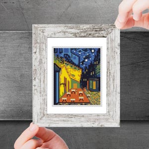 Puede incluir: Una impresión enmarcada de una pintura que representa una escena nocturna con una casa amarilla y un cielo azul estrellado. La casa tiene un techo rojo y una puerta amarilla. La pintura está en un marco blanco.