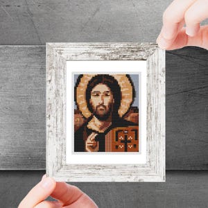 Puede incluir: Una obra de arte en punto de cruz enmarcada que representa una figura religiosa con un halo. La imagen presenta un diseño de punto de cruz detallado con hilos marrones, beige y negros. El marco es blanco con un acabado envejecido.