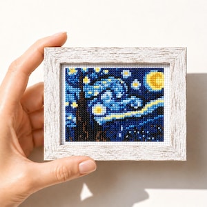 Op de afbeelding: Een ingelijste kruissteek van Van Goghs "Sterrennacht" met een wit, verweerde houten frame. Het kunstwerk toont een donkerblauwe lucht met wervelende gele en witte sterren, een gele maan en een donkere boom.