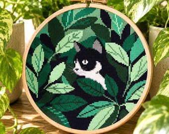 Könnte beinhalten: Ein runder Stickrahmen aus Holz mit einem Kreuzstichmuster, das eine schwarz-weiße Katze zeigt, die durch eine dichte Anordnung aus dunkel- und hellgrünen Blättern lugt. Die Katze hat weiße Abzeichen im Gesicht und an der Brust sowie sichtbare Schnurrhaare.