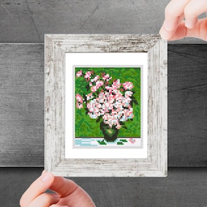 Può includere: Un ricamo a punto croce incorniciato che rappresenta un vaso pieno di fiori rosa su uno sfondo verde.