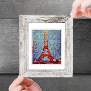 May include: A framed print of the Eiffel Tower in Paris, France. The tower is red and the background is a blue sky with white stars. The frame is white with a distressed wood finish.