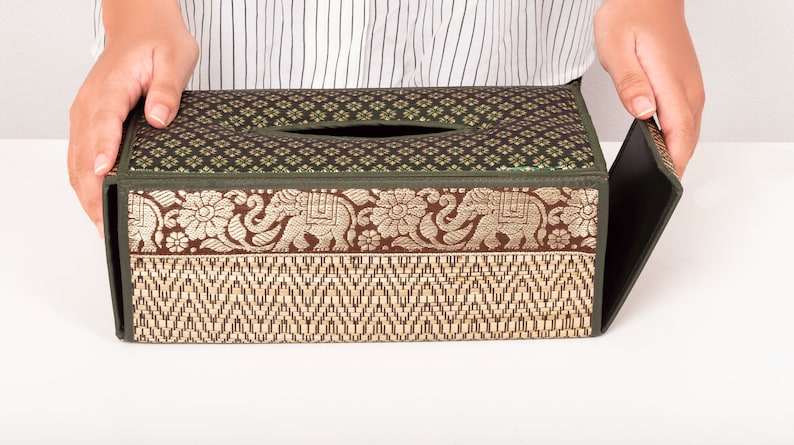 May include: A green and brown tissue box cover with a woven pattern and an elephant design. The cover is made of fabric and has a flap that opens to reveal the tissue box.
