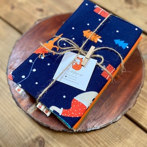 May include: A set of three fabric napkins with a blue background and a white polka dot pattern. The napkins feature a repeating pattern of orange foxes, pine trees, and presents. The napkins are tied together with twine and a wooden clothespin. The text on the tag reads "The Crafty Fox" and includes contact information.