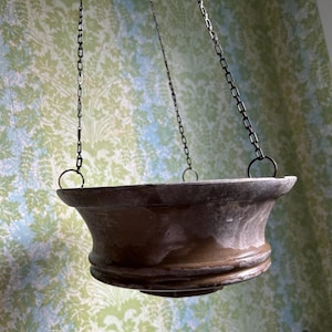 May include: A brown, hanging planter with a wide, flared bowl shape. The planter is suspended by dark metal chains attached to rings. The background features a wallpaper with a green and blue floral pattern.