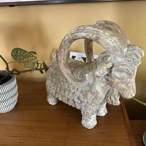 Op de afbeelding: Een verweerde, beige geitenfiguur met grote, gebogen hoorns. De sculptuur heeft een textuur en staat op een houten oppervlak. Een potplant is links zichtbaar.