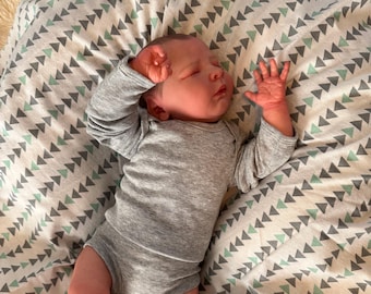 Könnte beinhalten: Ein schlafendes Baby in einem grauen Langarm-Strampler, das auf einer Decke mit einem geometrischen Muster aus grauen und grünen Dreiecken liegt. Die Hände des Babys sind an seinem Kopf erhoben.