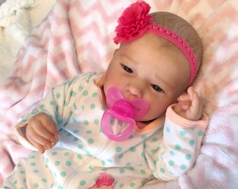 May include: A realistic baby doll wearing a white and green polka dot sleeper with a pink flower headband and a pink pacifier. The sleeper has the text "Cutest of them all" on the front. The doll is lying on a pink and white blanket.