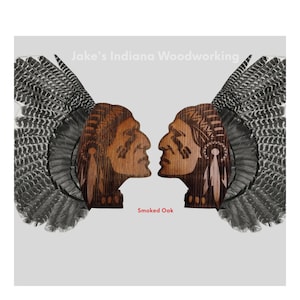 May include: Two wooden Native American profile carvings facing each other, with a turkey feather headdress on each side. The carvings are made of smoked oak wood. The text "Jake's Indiana Woodworking" is at the top of the image, and "Smoked Oak" is at the bottom.