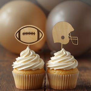 May include: Two cupcakes with white frosting, each topped with a laser-cut wooden topper. One topper is shaped like an American football, and the other is shaped like a football helmet. The toppers are mounted on thin wooden sticks.