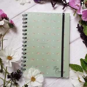 Può includere: Un quaderno a spirale verde con una chiusura a fascia elastica nera. La copertina presenta un motivo ripetuto di conigli marroni che corrono e cuori bianchi. Il testo "Love Country" è stampato in corsivo nero nell'angolo in basso a destra, con il nome "Sarah Reilly" stampato sotto in un carattere più piccolo.