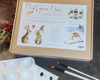 May include: A Christmas card kit in a cardboard box. The box includes paints, brushes, blank watercolor cards, envelopes, tracing paper, a pencil, an eraser, and a paint palette. The box has the text "Paint your Own Christmas Card Kit".