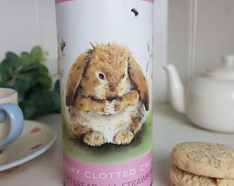 Può includere: Latta cilindrica decorativa con un'illustrazione di un coniglietto e la scritta "Luxury Clotted Cream Shortbread with Strawberry". La latta ha fasce rosa. Diversi biscotti shortbread sono impilati vicino a una teiera bianca.