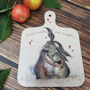 Op de afbeelding: Houten snijplank met handvat, met een illustratie van twee knuffelende hazen. De plank heeft de tekst "Fluffy cuddles and snuggles" en rode hartjes. Op de achtergrond zijn appels te zien.
