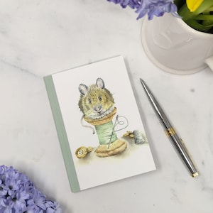 Può includere: Illustrazione ad acquerello su un taccuino con un topo che emerge da una bobina di filo verde. Il topo indossa un colletto a balze. Sono raffigurati anche un ago, un ditale e dei bottoni. Una penna d'argento e un vaso di fiori sono sullo sfondo.