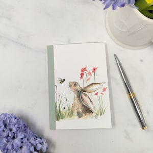 Può includere: Un'illustrazione ad acquerello di una lepre marrone con orecchie lunghe che guarda in alto a un bombo in volo. La lepre è circondata da fiori rosa ed erba verde. L'illustrazione è stampata su un piccolo taccuino rilegato in verde.