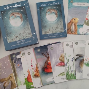 Può includere: Un set di carte oracle Wild Woodland con un libro guida. Le carte presentano illustrazioni ad acquerello di animali e piante del bosco. Il libro guida è blu con un titolo bianco e un design glitterato argento.