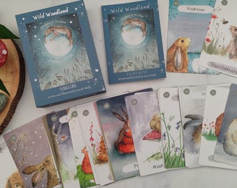 Pode incluir: Um conjunto de cartas de oráculo Wild Woodland com um guia. As cartas apresentam ilustrações em aquarela de animais e plantas da floresta. O guia é azul com um título branco e um design de glitter prateado.