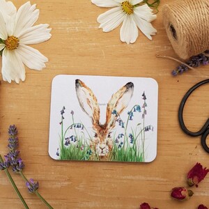May include: A rectangular coaster featuring a watercolor illustration of a brown hare peeking through green grass and bluebell flowers. The coaster has a white background and is surrounded by flowers and twine.