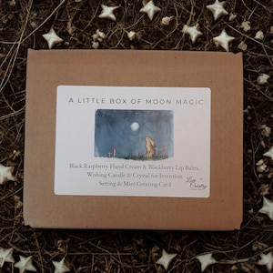 Op de afbeelding: Een bruine doos met een wit etiket met de tekst "A Little Box of Moon Magic". Het etiket toont een aquarel illustratie van een konijn onder een volle maan. De doos bevat handcrème, lippenbalsem, een kaars en een wenskaart.