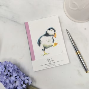 Könnte beinhalten: Ein kleines Notizbuch mit einem rosa Rücken, das eine Aquarell-Illustration eines Papageientauchers zeigt. Der Notizbuchumschlag trägt den Text "Love Country" und "Notebook" mit den Worten "96 linierte Seiten" und "Made in Britain".