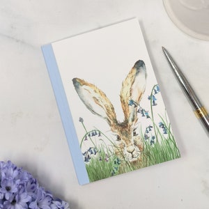 May include: A watercolour illustration of a brown rabbit with large ears peeking out from a field of bluebells. The rabbit is looking directly at the viewer with a curious expression.