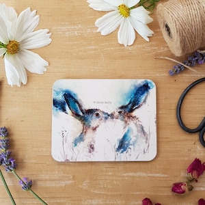 May include: Watercolor painting of two blue hares with their heads touching, set against a white background with a light blue wash. The painting is signed "Sarah Reilly" in the bottom right corner.