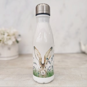Pode incluir: Garrafa de água de aço inoxidável branca com uma ilustração de um coelho castanho e branco rodeado de relva verde e flores azuis.